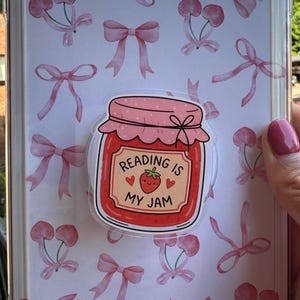 May include: A clear tablet case featuring a white background with a repeating pattern of light pink bows and cherries. A circular sticker is attached to the centre, depicting a red jam jar with a strawberry and the text Reading Is My Jam.