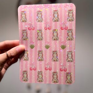 以下が含まれることがあります： ピンクのストライプの背景に、花の冠をかぶり、バスケットを持っているウサギのイラストが描かれた長方形のステッカーシート。 さくらんぼと星もデザインに含まれています。 ステッカーシートはキラキラした仕上がりです。