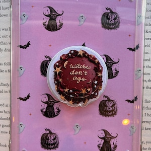 May include: A clear protective tablet case featuring a light purple insert decorated with black illustrations of cats in witch hats, pumpkins, ghosts, and bats. A circular phone grip with a dark red cake design and the text "witches don't age" is attached.