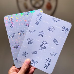 Puede incluir: Dos tarjetas rectangulares con fondo azul claro y un patrón de criaturas y objetos marinos. Las ilustraciones incluyen caballitos de mar, conchas, estrellas de mar, lazos, una langosta y rodajas de limón. Una tarjeta tiene un acabado holográfico.