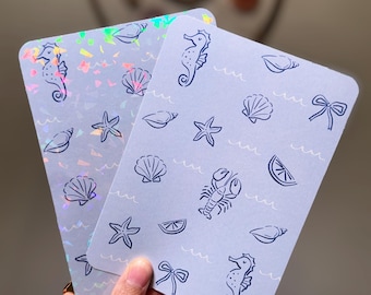 May include: Two rectangular cards with a pale blue background, featuring illustrations of seahorses, seashells, starfish, bows, a lobster, and lemon slices. One card has a holographic finish, creating a shimmering effect.