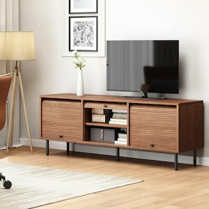 May include: A mid-century modern style TV stand in a warm brown finish. The stand features two sliding doors with a slatted design, a central open shelf, and black metal legs. A large flat-screen TV sits on top.