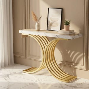 May include: A modern console table with a marble top and gold-colored, curved metal base. The table features a rectangular top with a light-colored marble pattern. The base has a unique, arched design. A framed artwork, small plant, and decorative items are on the table.