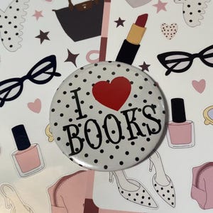 May include: A white button with black polka dots and the text "I ❤️ BOOKS" in black, with a red heart. The button is surrounded by stickers of lipstick, eyeglasses, nail polish, and other fashion-themed items.