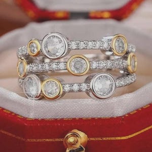 Puede incluir: Un anillo de tres niveles con una banda plateada y detalles dorados. El anillo presenta múltiples piedras preciosas redondas y transparentes engastadas en un bisel. El anillo se exhibe en una caja de joyería roja con forro de satén blanco.