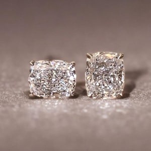 May include: A pair of square-cut diamond stud earrings. The diamonds are set in a silver or white gold setting. The earrings are displayed on a gray surface.