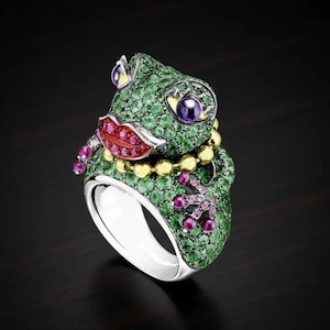 May include: A silver ring shaped like a frog, featuring a body covered in small green stones, large purple eyes, bright red lips, and a gold-toned beaded necklace. The frog's feet are accented with small pink stones, and the band is polished silver.