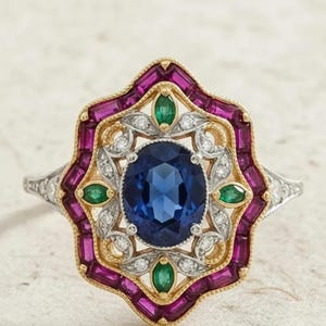 Peut inclure: Une bague ornée d'une grande pierre bleue ovale au centre, entourée d'une monture en or et argent. La bague est ornée de petits accents d'émeraudes et de rubis, créant un design d'inspiration vintage.