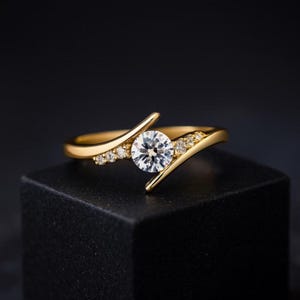 Puede incluir: Un anillo de oro con una gema grande, redonda y transparente como punto focal. El anillo presenta un diseño de caña dividida con piedras de acento más pequeñas. El anillo se exhibe sobre una superficie negra sobre un fondo oscuro.