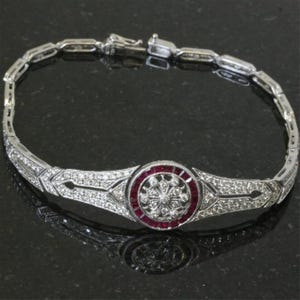 May include: A silver bracelet with a central circular design featuring a ruby-coloured centre and a diamond-like pattern. The bracelet has a delicate, intricate design with small, sparkling stones throughout.
