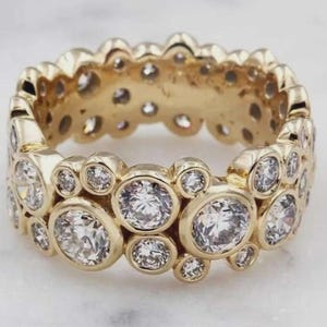 Puede incluir: Un anillo dorado con múltiples piedras preciosas redondas y transparentes. El anillo presenta un diseño de doble banda, con las piedras engastadas en un estilo de bisel. El anillo es probablemente de oro y es una joya.