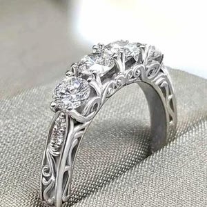 May include: A silver ring with three large, round, clear gemstones set in a decorative band. The band features intricate filigree designs and smaller accent stones, creating a classic and elegant look. The ring is displayed on a textured, light gray surface.