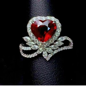 May include: A heart-shaped, red gemstone ring with a halo of small, clear stones. The ring's band features a floral design with additional clear stones, set against a black background. A statement piece of jewelry.
