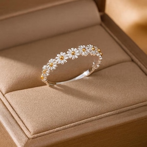 May include: A silver ring featuring a daisy flower design. The ring has small white petals with gold-colored centers. The ring is displayed in a brown jewelry box.