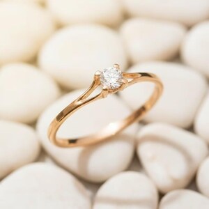 Op de afbeelding: Een goudkleurige ring met een enkele, heldere edelsteen, gezet in een vier-klauwzetting. De ringband splitst zich in twee delicate strengen aan weerszijden van de edelsteen. De ring ligt op witte stenen.