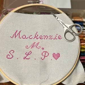 May include: A cross-stitch project in a wooden embroidery hoop. The project features the name "Mackenzie" and initials, all stitched in pink thread. A pair of small silver scissors rests on the fabric, ready for trimming. The craft project is on a white fabric.