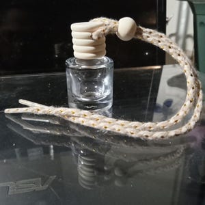 May include: A small, clear glass bottle with a light-colored wooden cap and a decorative rope. The rope is beige with gold accents and a small wooden bead. The bottle is likely an essential oil diffuser or air freshener.