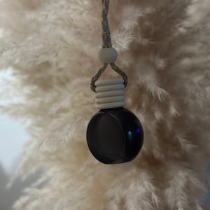 May include: A black, spherical car air freshener with a gray and gold braided cord. The air freshener has a white, ribbed base and a white bead. The background is a fluffy, cream-colored texture.