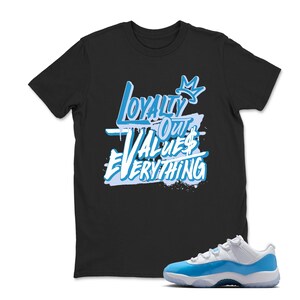 Puede incluir: Camiseta negra con el texto "Loyalty Out Values Everything" en azul claro y blanco. Un par de zapatillas blancas y azul claro est&aacute; al lado de la camiseta.