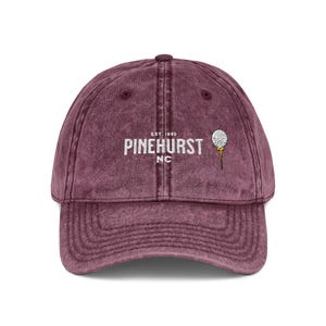 May include: A maroon baseball cap with a curved brim. The cap features the text "PINEHURST NC" in white, with "EST. 1895" above it. A golf ball on a tee is embroidered on the right side. The cap is made of a soft, textured material.