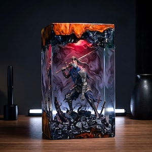 May include: A detailed resin sculpture of a warrior figure, holding a sword, encased in a rectangular block. The sculpture features a dark color palette with red and orange accents. The warrior stands on a rocky base, with additional swords embedded.