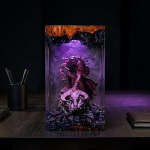 Puede incluir: Escultura rectangular de resina que representa a un guerrero arrodillado sobre una calavera. La escultura está encerrada en resina transparente con un efecto ahumado púrpura y blanco, y una base de madera oscura. El guerrero sostiene una espada.