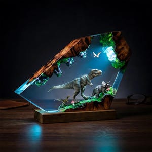 Puede incluir: Lámpara temática de dinosaurios con un T-Rex, Triceratops y otras criaturas prehistóricas en una estructura de resina transparente. La lámpara tiene una base de madera y una luz azul, creando una exhibición cautivadora.