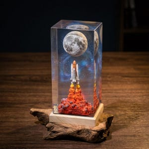 May include: A clear resin block depicting a space shuttle launching towards a full moon against a starry backdrop. The base is a wooden block, and the piece is mounted on a piece of driftwood.