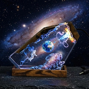 May include: A decorative space-themed lamp featuring an astronaut, Earth, and a galaxy design. The lamp is encased in a clear, angled resin with a wooden base. The background showcases a vibrant depiction of the Milky Way.