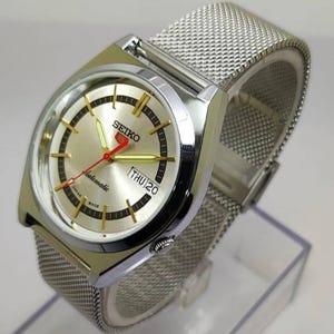 May include: A silver-toned Seiko automatic wristwatch with a mesh band. The watch face is gold with black and gold markers, a red second hand, and the day displayed. The watch is marked "automatic" and "Japan Made".