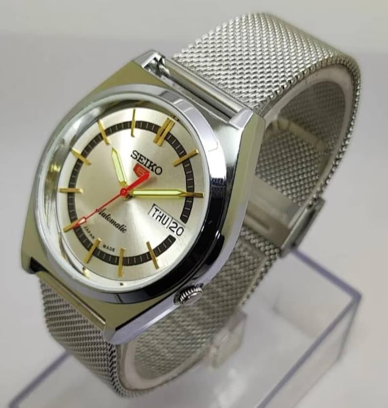 May include: A silver-toned Seiko automatic wristwatch with a mesh band. The watch face is gold with black and gold markers, a red second hand, and the day displayed. The watch is marked "automatic" and "Japan Made".