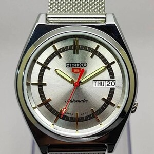May include: Silver-toned Seiko 5 automatic watch with a mesh band. The watch face is silver with black and gold accents, including the day and date display. The watch reads "Automatic" and "Japan Made".