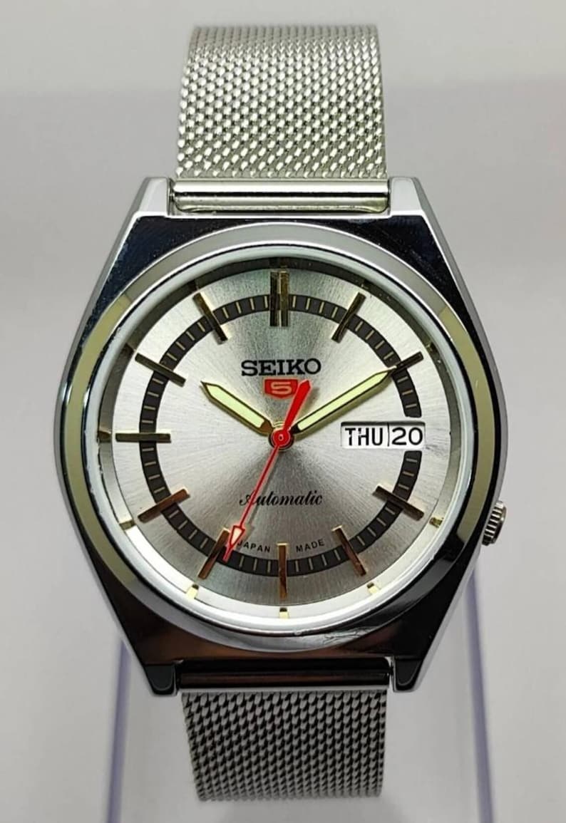 May include: Silver-toned Seiko 5 automatic watch with a mesh band. The watch face is silver with black and gold accents, including the day and date display. The watch reads "Automatic" and "Japan Made".