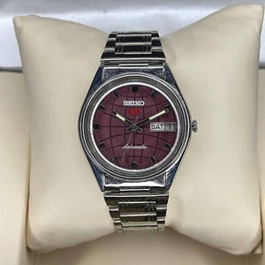 Può includere: Un orologio Seiko 5 Automatic vintage con un quadrante bordeaux con motivo a griglia. L'orologio ha un cinturino e una cassa in acciaio inossidabile color argento. Il giorno è impostato su sabato e l'orologio è esposto su un cuscino color crema.