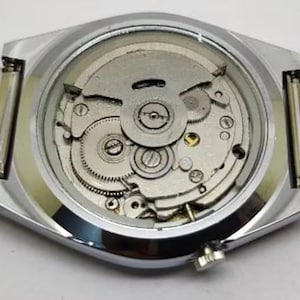 May include: Close-up of the back of a silver wristwatch with a mesh band. The watch case is silver and octagonal, with a clear back revealing the intricate mechanical movement. The band is a fine, woven silver mesh.