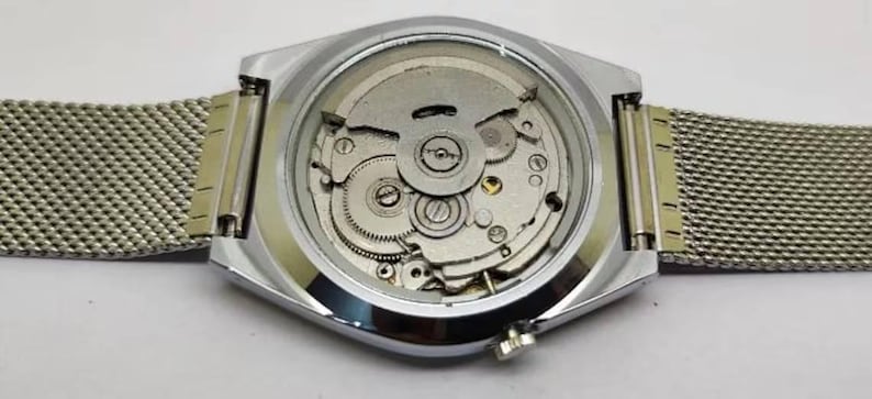 May include: Close-up of the back of a silver wristwatch with a mesh band. The watch case is silver and octagonal, with a clear back revealing the intricate mechanical movement. The band is a fine, woven silver mesh.