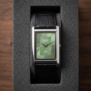 Puede incluir: Un reloj Seiko Quartz rectangular con esfera verde y caja plateada, presentado en una caja gris. El reloj presenta números romanos y una correa de cuero negro.