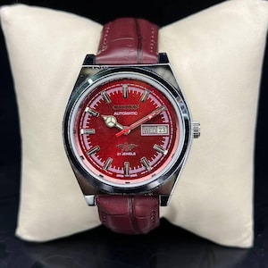 Puede incluir: Un reloj de pulsera automático Citizen con esfera roja y correa de cuero burdeos. La esfera muestra el día y la fecha, con el texto "WED 23". El reloj tiene una caja plateada y está sobre un cojín blanco.