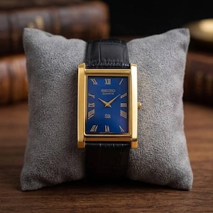 May include: A rectangular gold-toned wristwatch with a blue face and black leather strap. The watch face features Roman numerals and gold hands. The watch is displayed on a grey pillow against a wooden background. The watch is a Seiko Quartz.