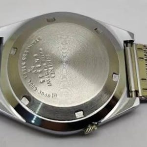 May include: A silver-toned wristwatch with a mesh band, viewed from the back. The watch face is a geometric shape, and the back casing has engraved text. The band is a fine, woven metal. The watch is on a white surface.
