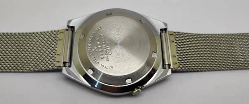 May include: A silver-toned wristwatch with a mesh band, viewed from the back. The watch face is a geometric shape, and the back casing has engraved text. The band is a fine, woven metal. The watch is on a white surface.