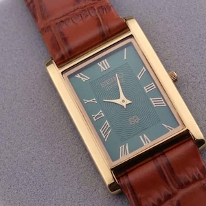 May include: A rectangular gold-toned wristwatch with a teal face and Roman numeral hour markers. The watch has gold hands and a brown leather band with a crocodile pattern. The watch is displayed on a gray surface.