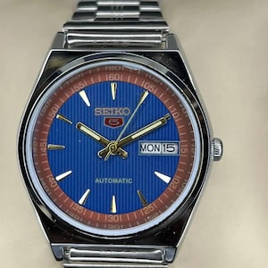 May include: A silver Seiko 5 automatic wristwatch with a blue dial and a brown outer ring. The watch features gold hands and markers, and a day-date display. The watchband is silver.