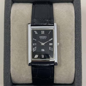 May include: A rectangular Seiko Quartz watch with a black face and silver-tone case. The watch features Roman numeral hour markers and a black leather band. The watch is presented in a gray box.