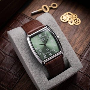 Pode incluir: Um relógio Seiko Quartz com mostrador verde retangular e caixa prateada, com algarismos romanos. O relógio tem uma pulseira de couro castanho com padrão de crocodilo. O relógio é exibido em uma almofada cinza, com uma chave e engrenagens no fundo.