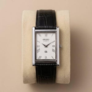 Puede incluir: Un reloj Seiko Quartz rectangular con caja plateada y correa de cuero negro. La esfera blanca presenta números romanos y el logotipo de Seiko. El reloj se muestra sobre un cojín beige.