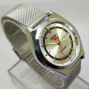 May include: Silver-toned Seiko 5 automatic watch with a mesh band. The watch face is silver with gold-toned markers and hands, including a red second hand. The day and date are displayed at the 3 o'clock position. The watch is marked "SEIKO" and "Automatic".