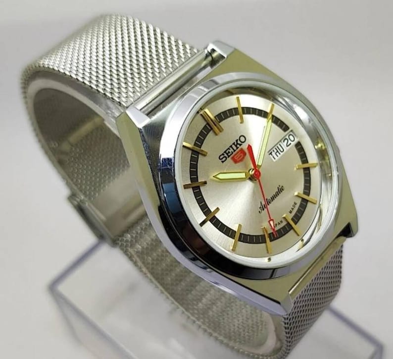 May include: Silver-toned Seiko 5 automatic watch with a mesh band. The watch face is silver with gold-toned markers and hands, including a red second hand. The day and date are displayed at the 3 o'clock position. The watch is marked "SEIKO" and "Automatic".