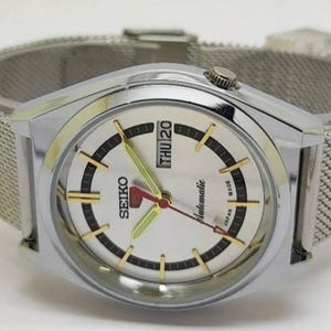 May include: A silver-toned Seiko automatic wristwatch with a mesh band. The watch face is white with black and gold accents, and displays the day and date. The hands are red and green.