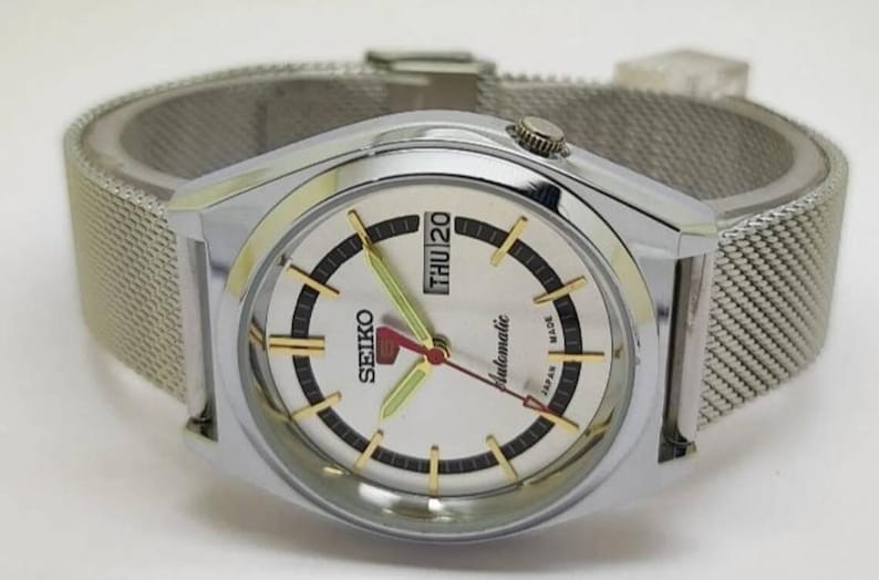 May include: A silver-toned Seiko automatic wristwatch with a mesh band. The watch face is white with black and gold accents, and displays the day and date. The hands are red and green.
